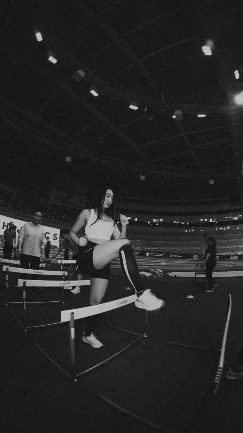 A woman in a sports outfit is stepping over a hurdle on a track, with additional hurdles and other people visible in the background, inside of a stadium.