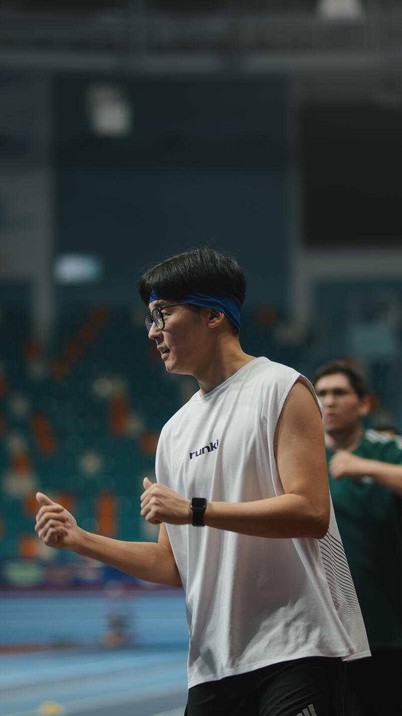 A man with glasses and a blue headband is wearing a white tank top with the word "runki" printed on it, and he is on an indoor track.
