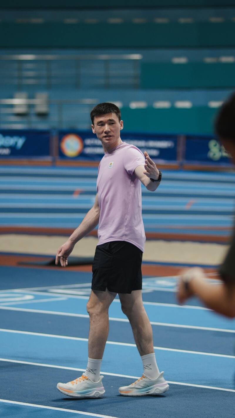 A man stands on a blue track wearing a lilac t-shirt, black shorts, white socks, and running shoes, gesturing with his right hand.