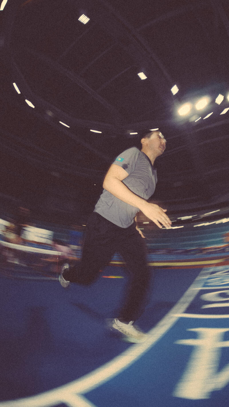 A man in a grey t-shirt with the Kazakhstan flag on the sleeve and black pants is running on a blue track, with motion blur indicating speed.