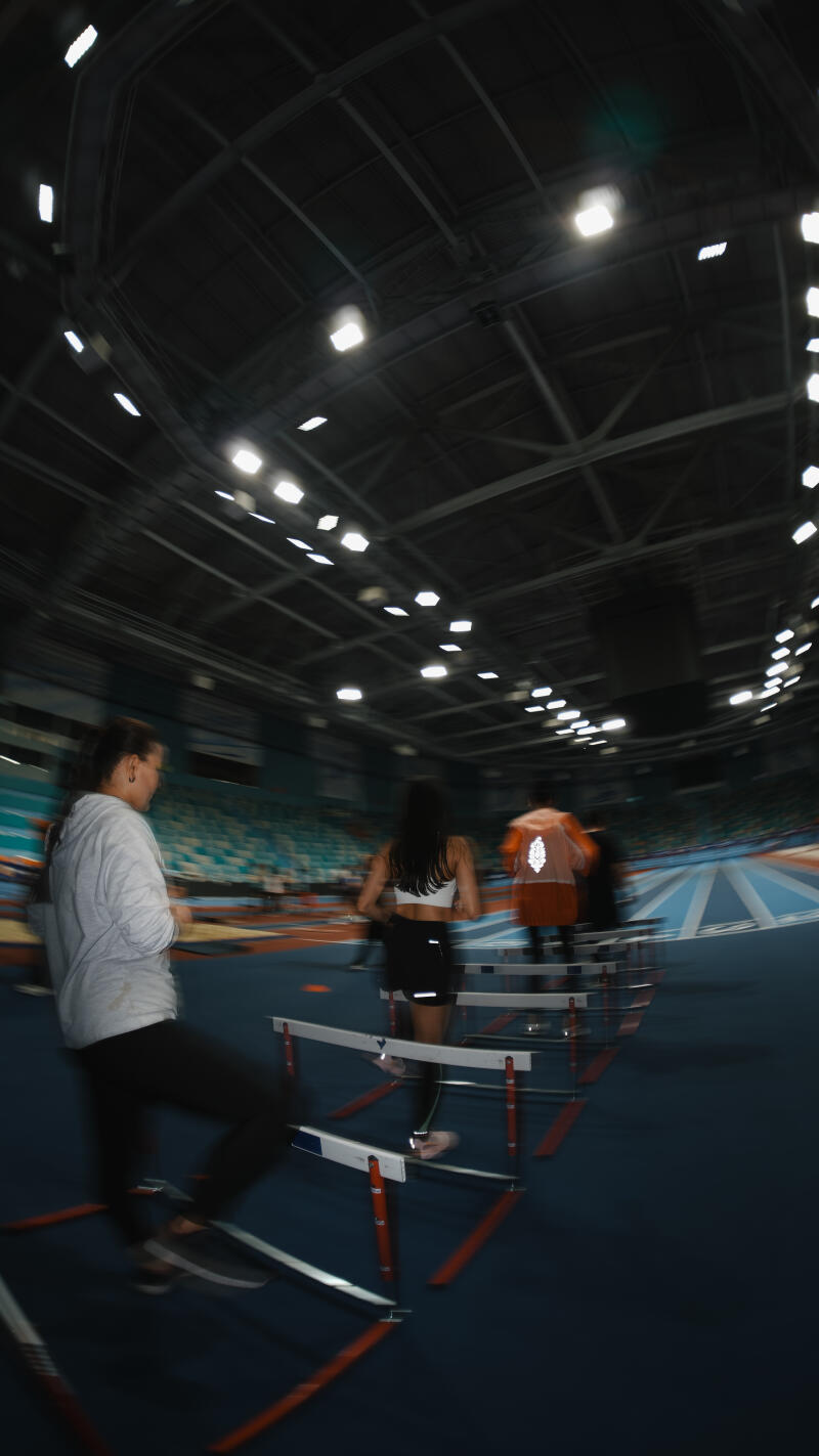 The image shows several athletes training on a track, jumping over hurdles under the bright lights of an indoor stadium.