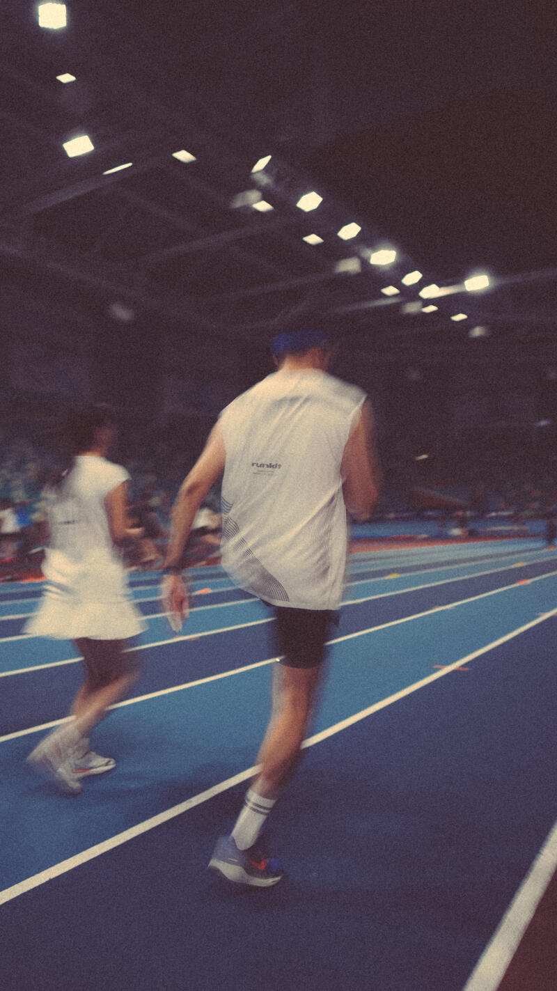 The image shows two runners in motion on a blue track with white lines, under bright, hexagonal lights. The runner in the foreground wears a white sleeveless shirt with the text 'runkdz' and black shorts.