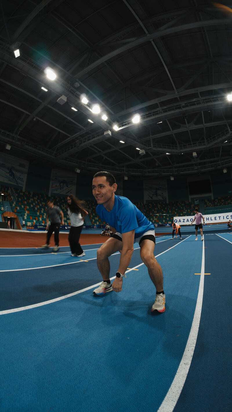 A man in a blue shirt and athletic shorts is posed on a blue running track, with other people and stadium seating visible in the background.