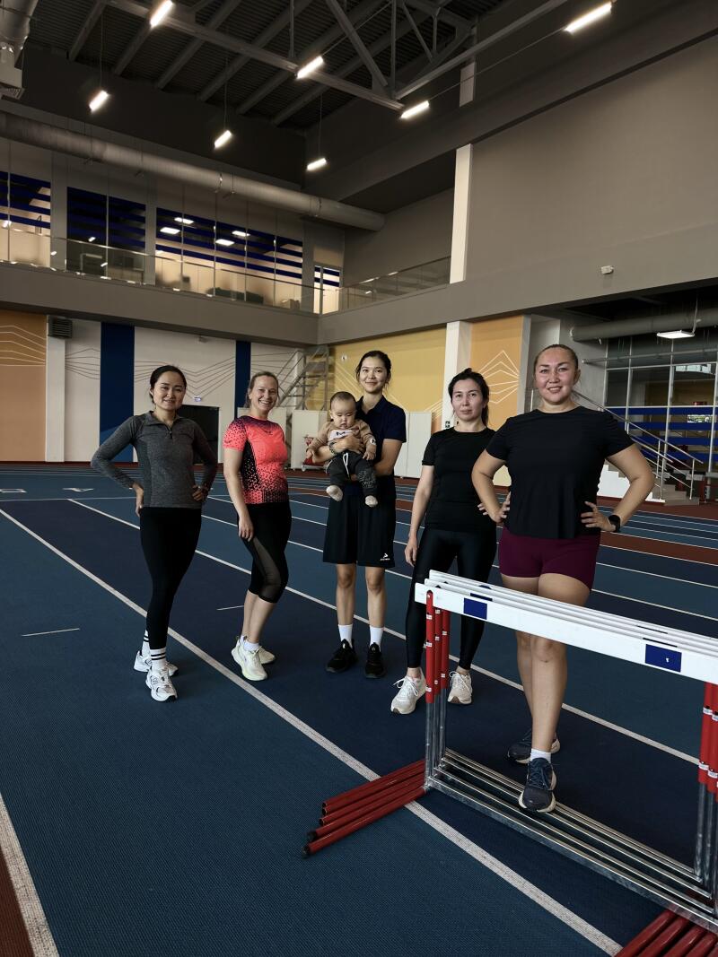 Five women stand on a blue running track, one holding a baby, with a hurdle in the foreground.