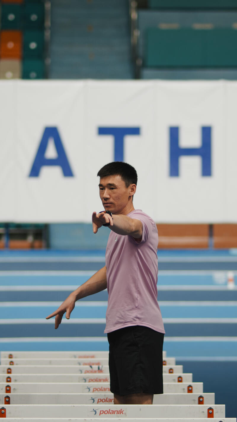 A man in a pink shirt and black shorts is standing on a blue track in front of hurdles, with the word 'ATH' visible in the background.