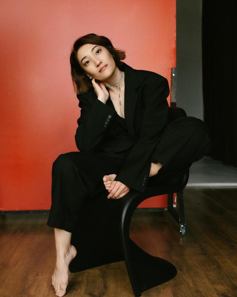 A woman with short brown hair is sitting barefoot on a black chair wearing a black suit and a silver necklace, posing in front of a red wall.