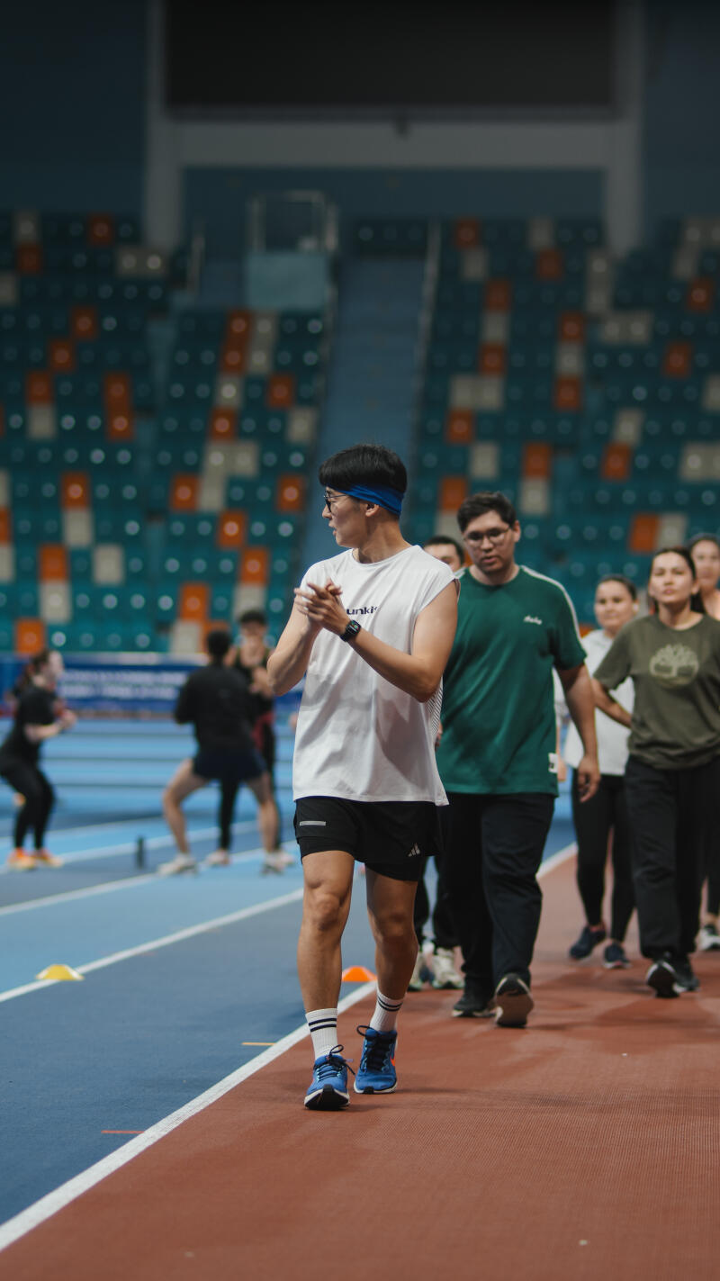 A group of people is walking on a running track in a stadium; one man in the foreground is wearing a white t-shirt, black shorts, blue shoes, and a blue headband.