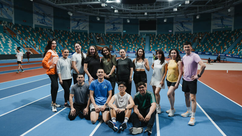 A group of approximately 15 people is posing on an indoor running track, with seating visible in the background.