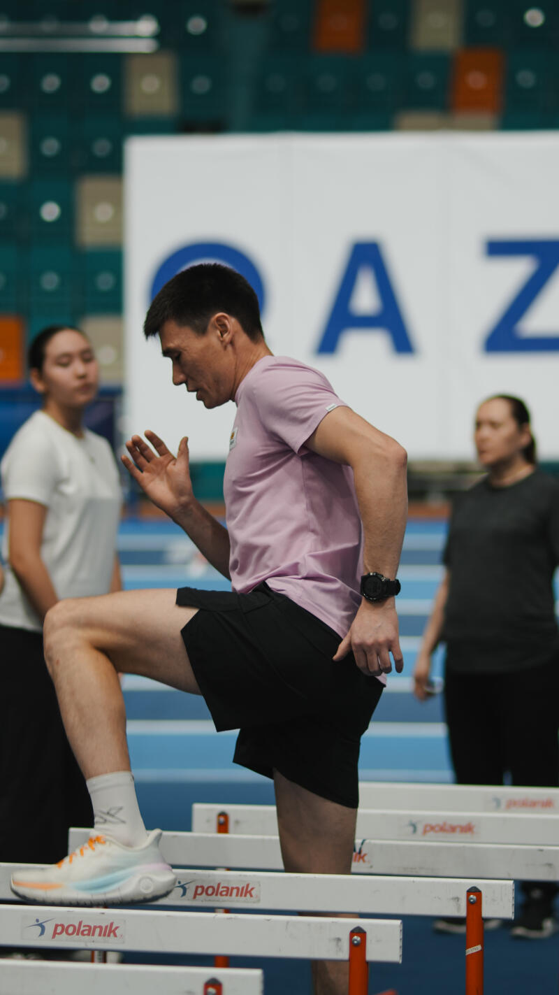 A man in a pink shirt and black shorts steps over hurdles, with two women in the background and the word "polanik" visible on the hurdles.