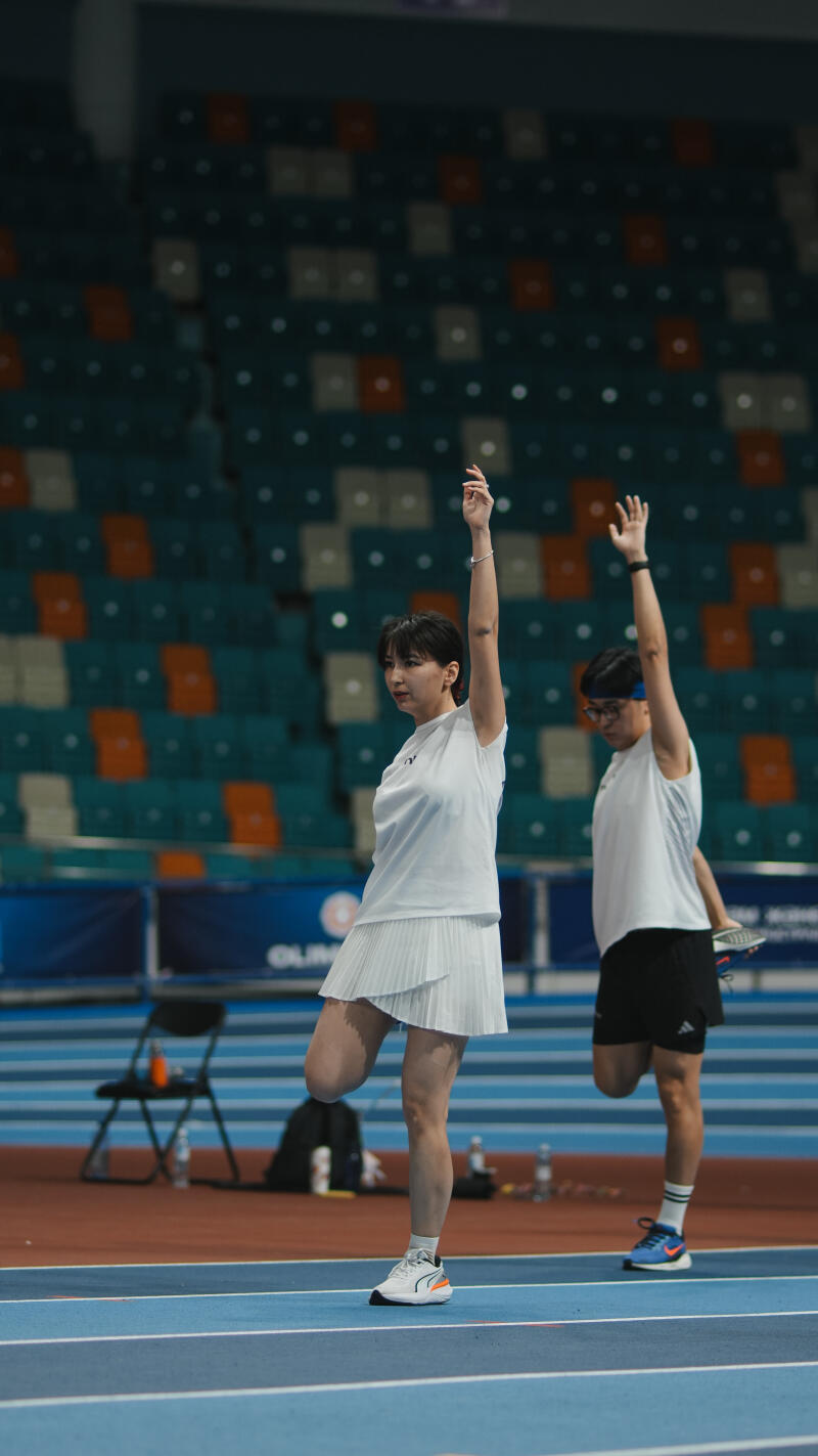 Two athletes are stretching on a blue running track with raised arms, wearing white shirts and dark shorts or skirts, in front of stadium seating.