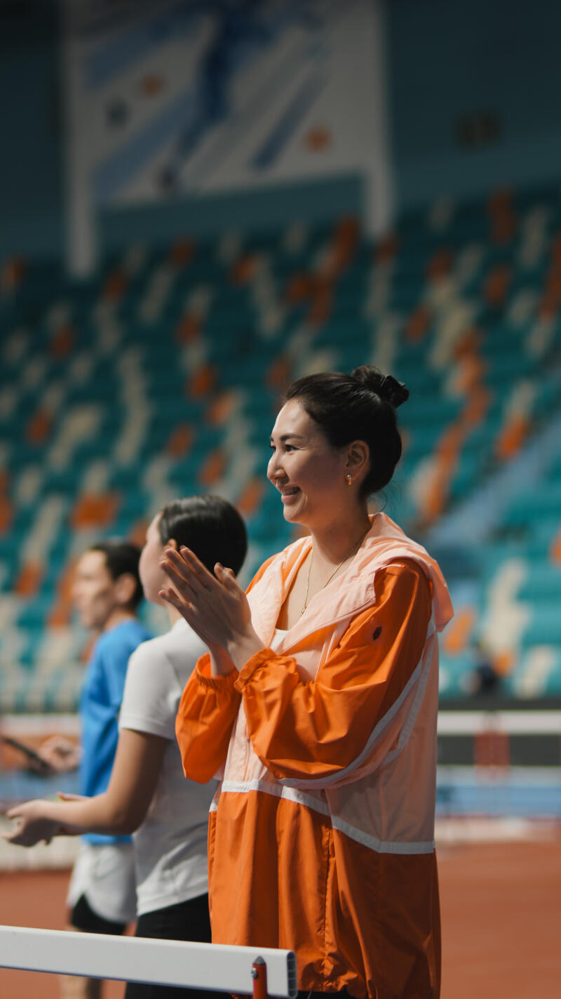 A smiling woman in an orange jacket is clapping, with other people and stadium seating visible in the background.