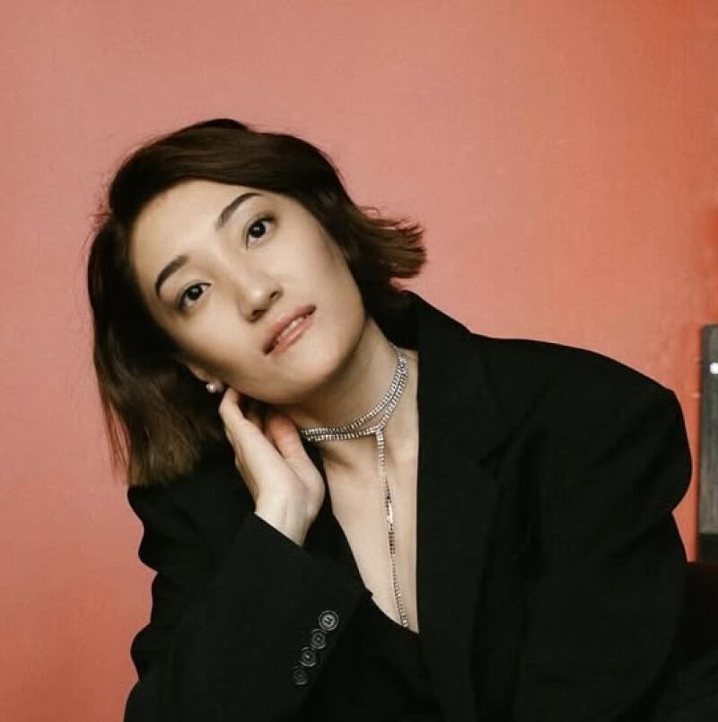 A woman with short brown hair poses in a black blazer with a silver necklace against a peach background.