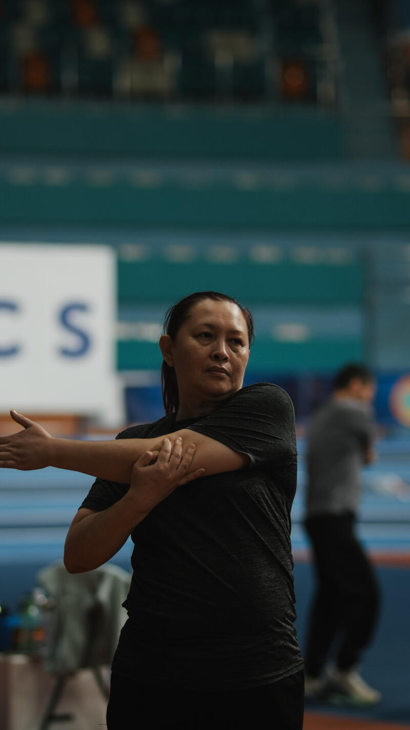 A woman in a gray shirt is stretching her arm while standing in a gymnasium with the partial text "CS" visible in the background.
