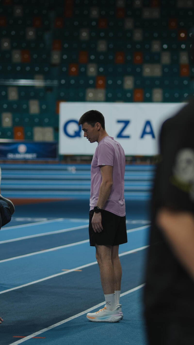 A man in a pink shirt and black shorts stands on a blue track with the text "Qazaqstan" in the background.