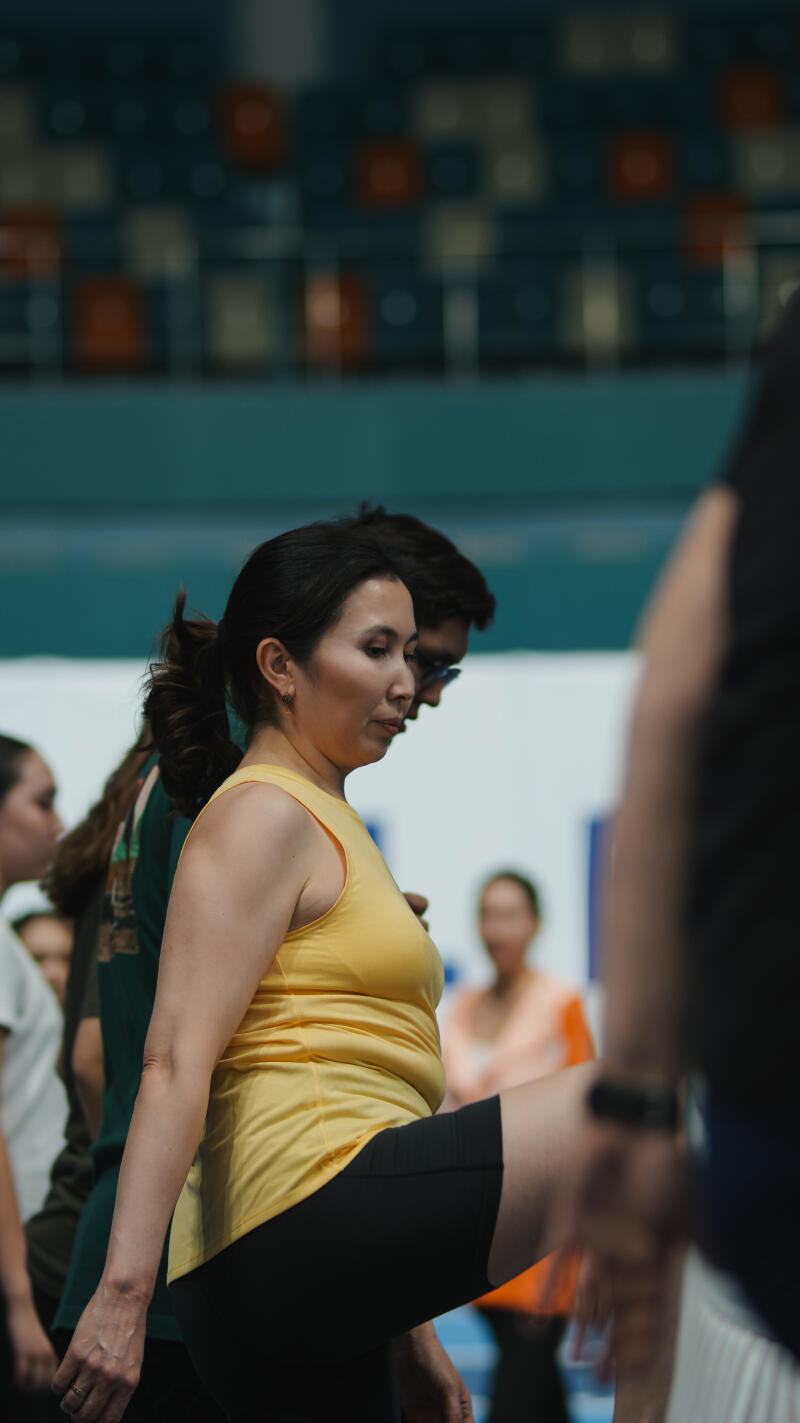 A woman in a yellow tank top and black shorts is seen in a gymnasium, surrounded by other people.