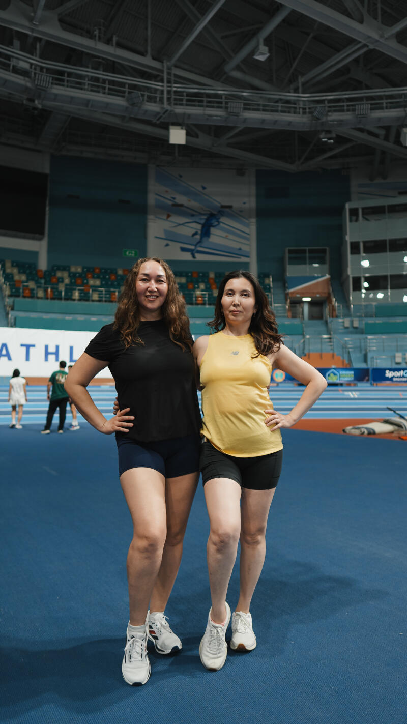 Two women stand side-by-side on a blue track, one in a black shirt and blue shorts, and the other in a yellow tank top and black shorts, both wearing sneakers.