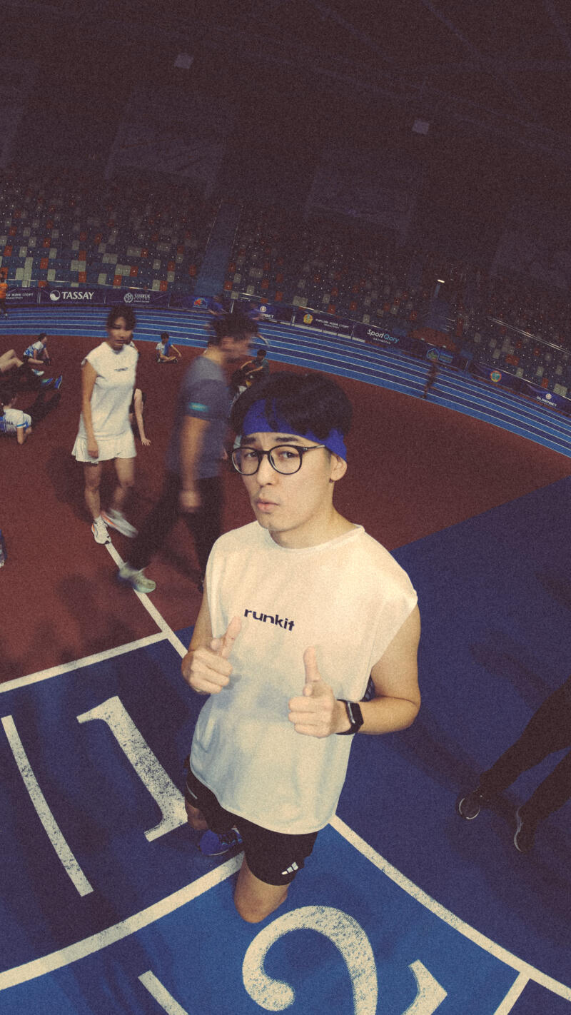 A man wearing glasses and a headband gives two thumbs up while standing on a blue track marked with the number '1'. He is wearing a 'runkit' t-shirt, black shorts, and a smartwatch, with an audience and other athletes visible in the background.