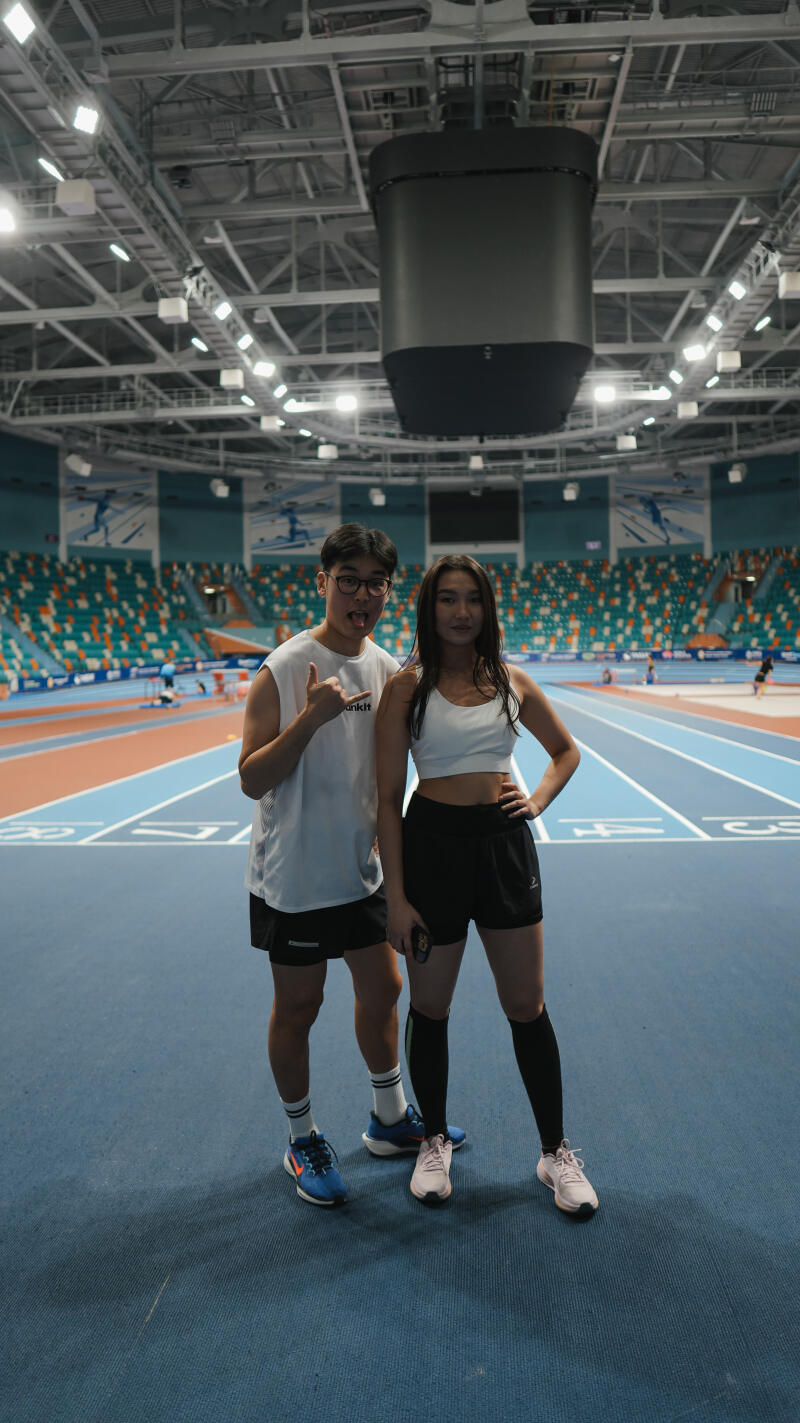 A man and a woman are standing on a blue indoor track, dressed in athletic wear, with stadium seating and a large electronic screen in the background.