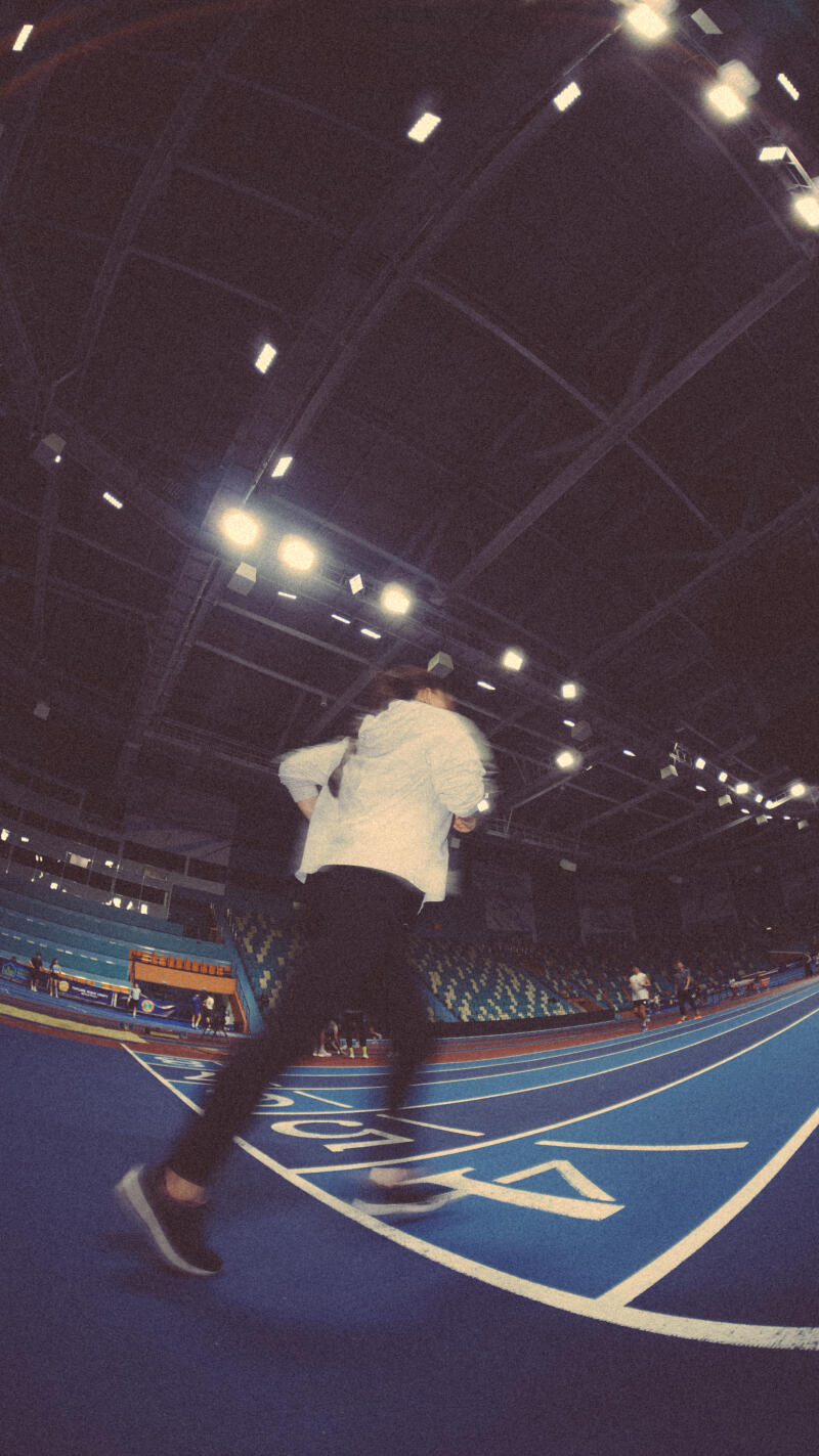 A blurry image of a person jogging on a blue track in an indoor stadium with stadium seating and overhead lighting.