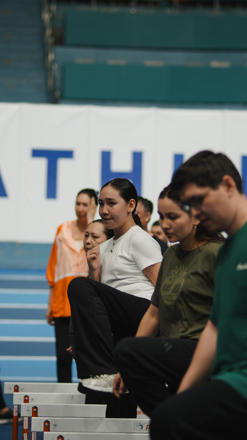 A group of people are sitting on hurdles in an indoor stadium; a white banner with large blue letters is in the background.