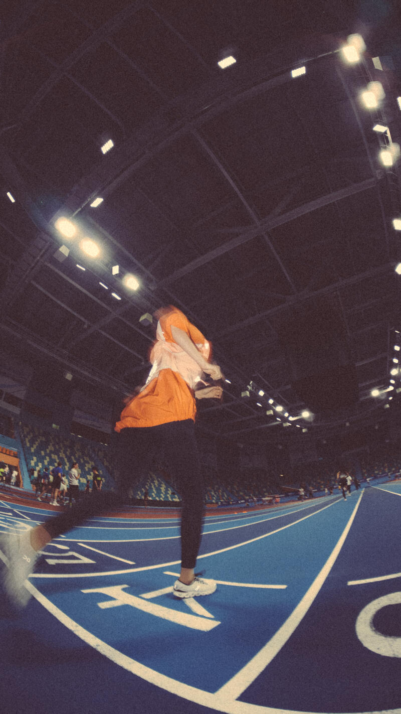 A blurry image shows a person in an orange shirt and dark pants running on a blue track with white lines in a stadium; a crowd is visible in the background and the ceiling is lined with lights.
