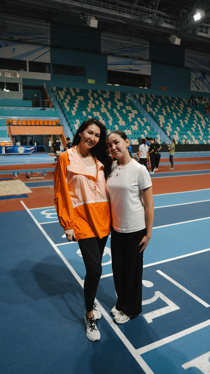 Two women stand on a blue track wearing athletic clothing, with stadium seating in the background.
