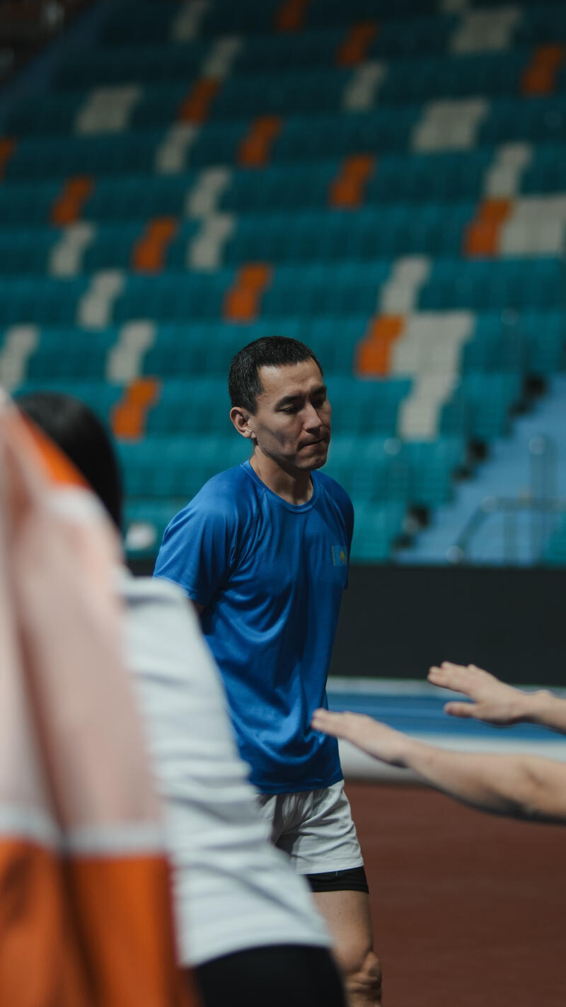 A man in a blue shirt with a Kazakhstan flag patch stands on a track field with other people and seating in the background.