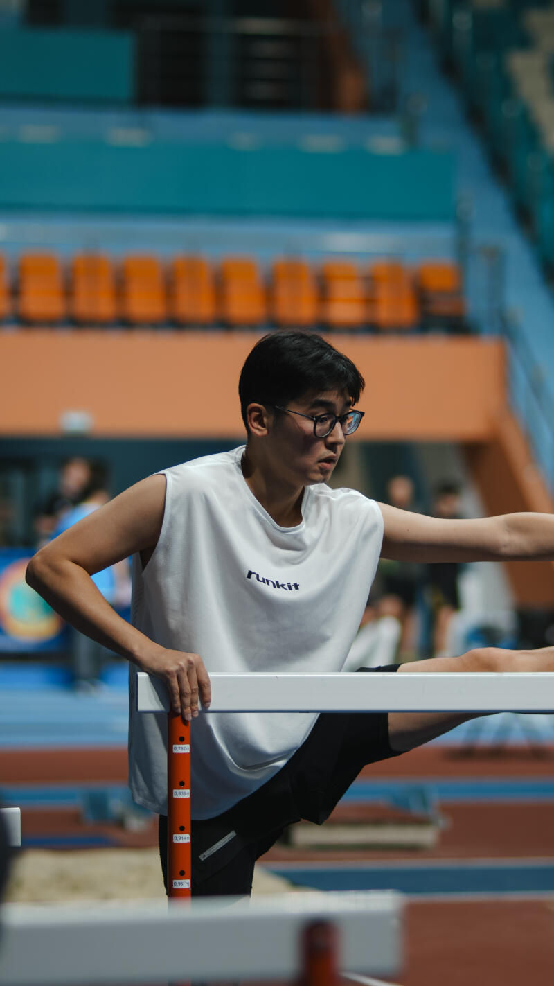 A young man wearing a white tank top with the word "funkit" on it and glasses is stretching his leg on a hurdle.