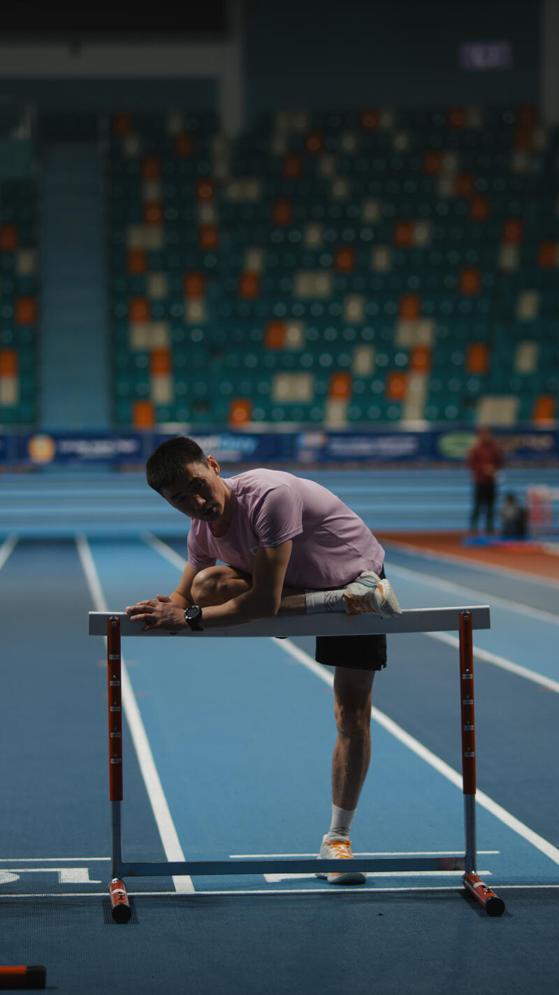 A man in a pink shirt and black shorts is stretching on a hurdle on a blue track.