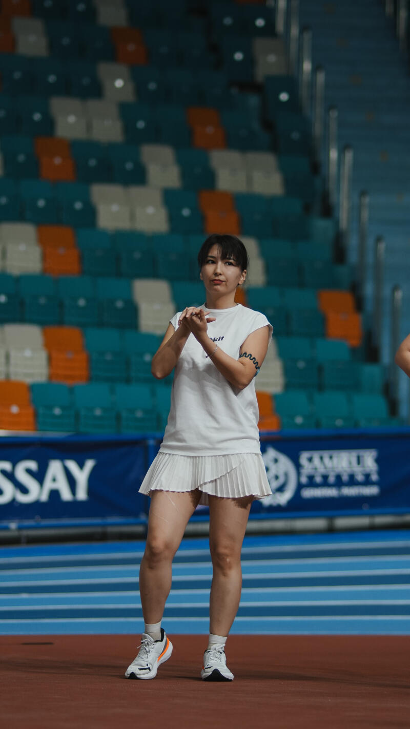 A woman in a white shirt and skirt stands on a red track in front of stadium seating, rubbing her hands together.