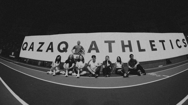 A group of nine people are sitting on a track in front of a banner that reads "QAZAQ ATHLETICS."