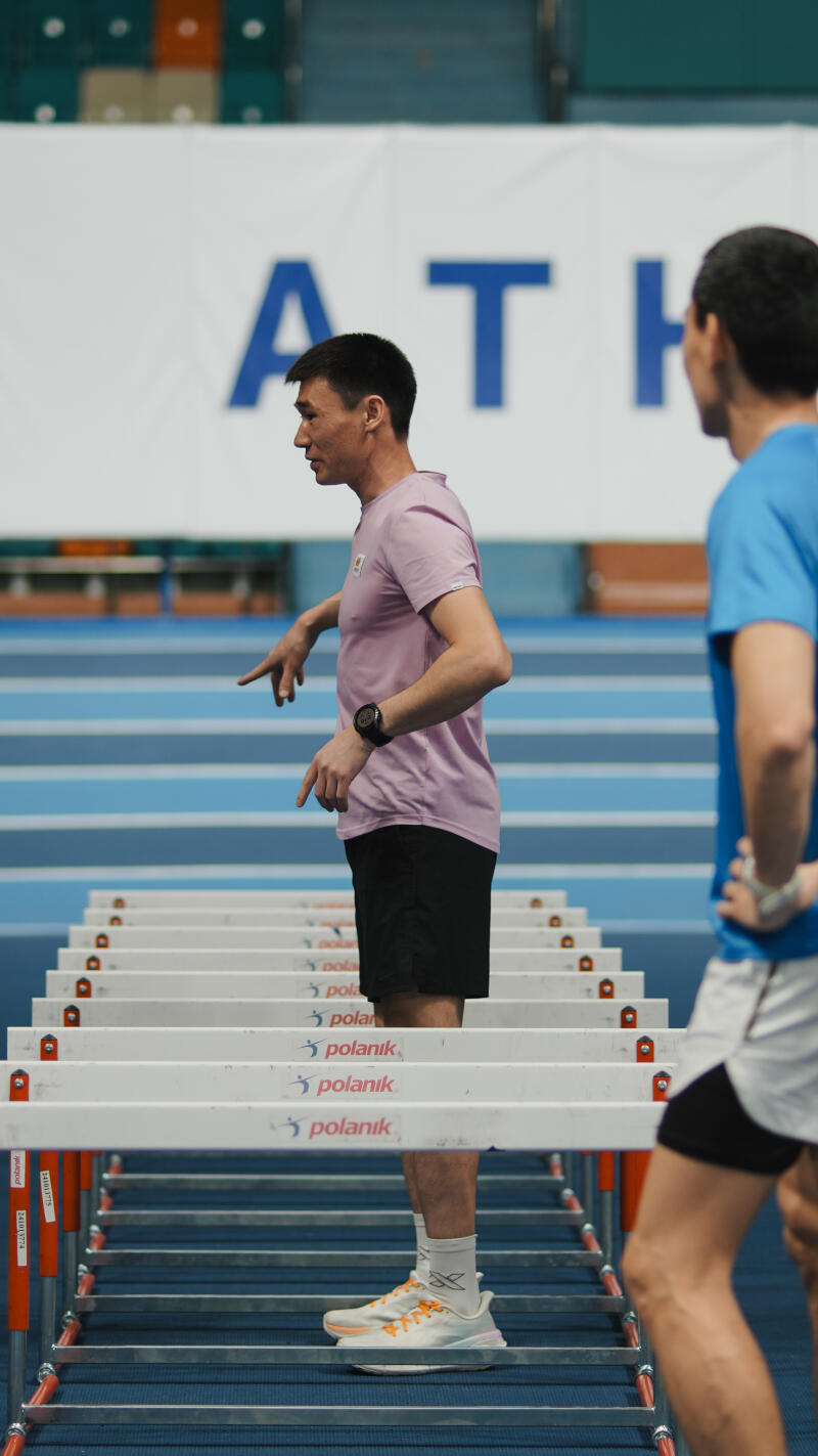 A man in a pink t-shirt and black shorts is standing on a blue track next to hurdles, gesturing with his hands, while another man in a blue t-shirt is standing to his right; the word 'ATH' is visible in the background.