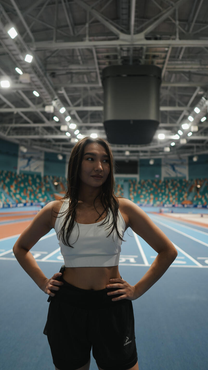 A woman in a white sports bra and black shorts with the 'Demix' logo stands with her hands on her hips, on a blue running track inside a large indoor stadium.
