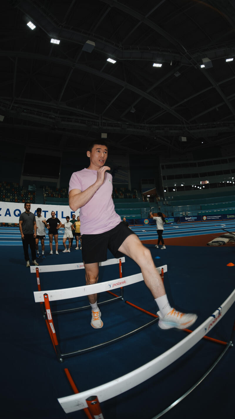 In an indoor stadium setting, an athlete in a pink shirt and black shorts is stepping over a hurdle, with other people standing in the background.