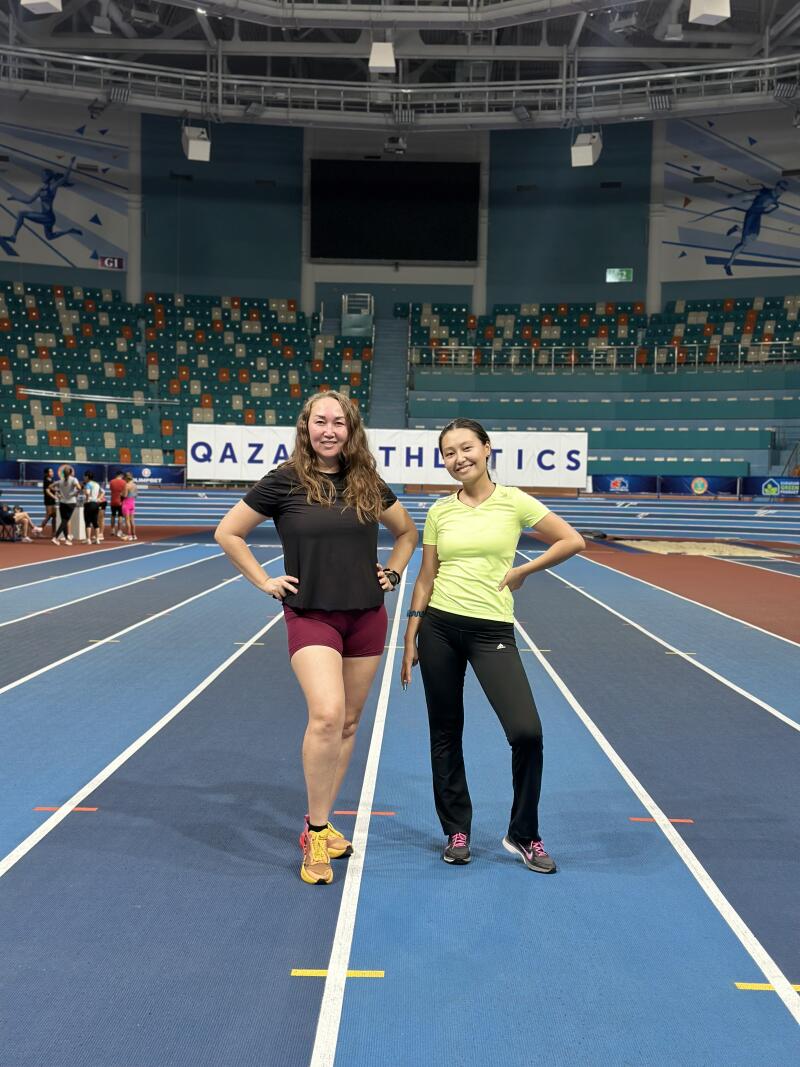 Two women are standing on a blue running track in front of stadium seating, with "QAZA ATHLETICS" written on a sign in the background.