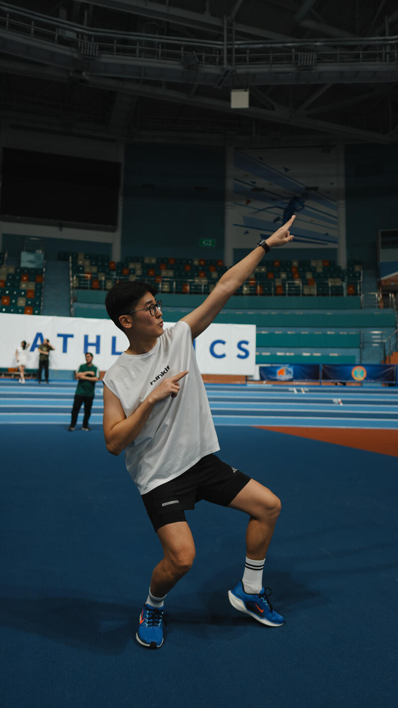 An Asian man wearing glasses, a white tank top, and black shorts stands on a blue track pointing up and to the side with both hands. Behind him is a banner that reads "ATHLETICS."