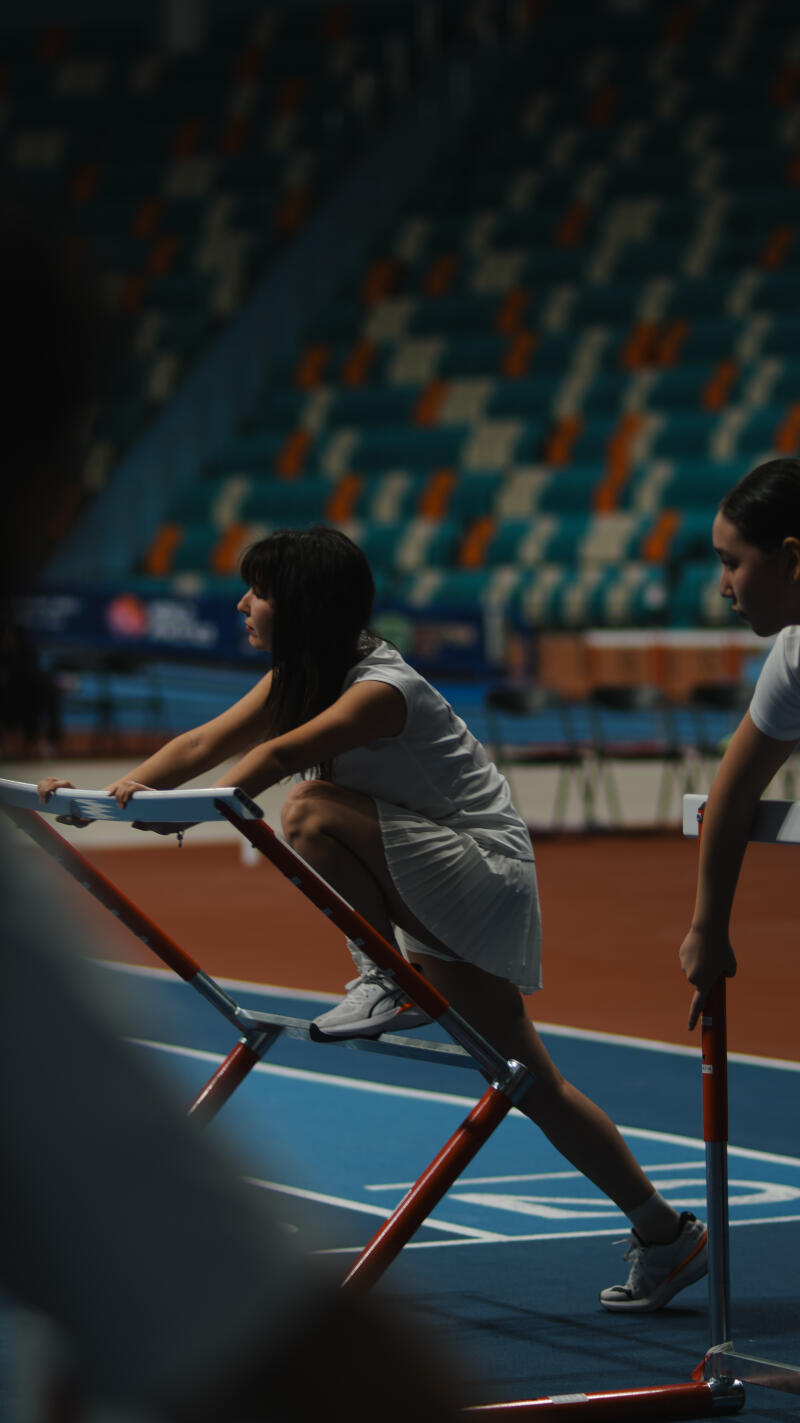 Two women in athletic attire are on a blue and red track with hurdles; one woman is squatting, holding onto a hurdle, while the other stands, holding a hurdle.