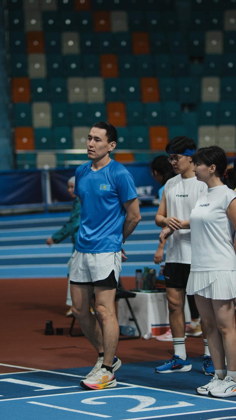 A man in a blue shirt with the Kazakhstan flag is standing on a running track next to two people wearing white shirts with the word "runkit" on them.