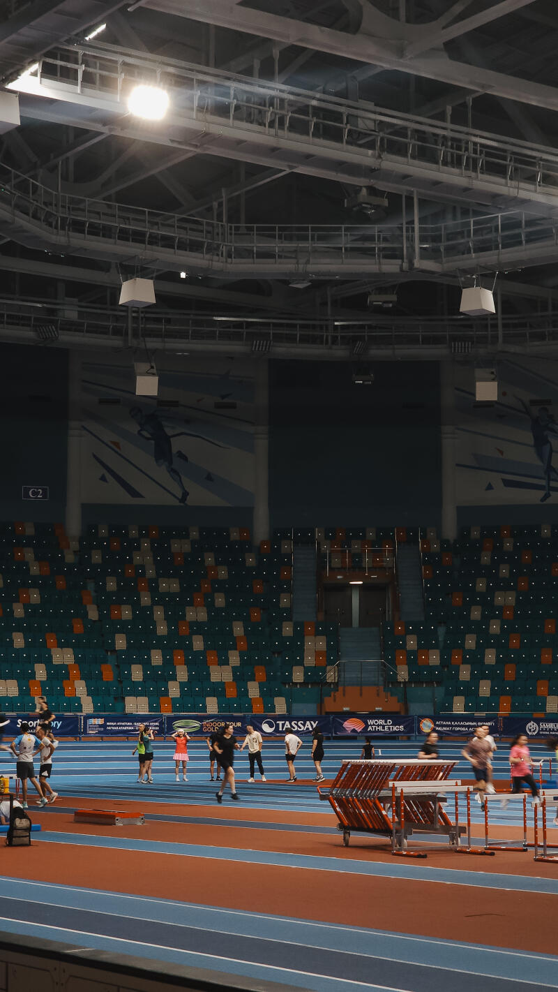 A wide shot shows an indoor track and field arena with athletes training on a blue track, surrounded by rows of orange and green seats, under a complex ceiling structure with bright lights.
