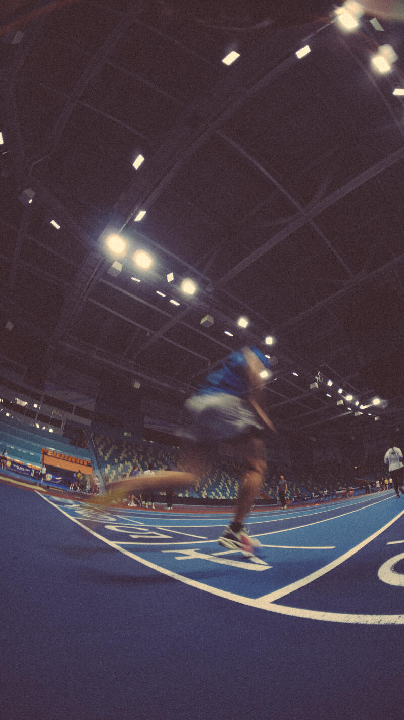 A blurry photo of a person running on a blue track in a stadium, with stadium seating and ceiling lights visible in the background.