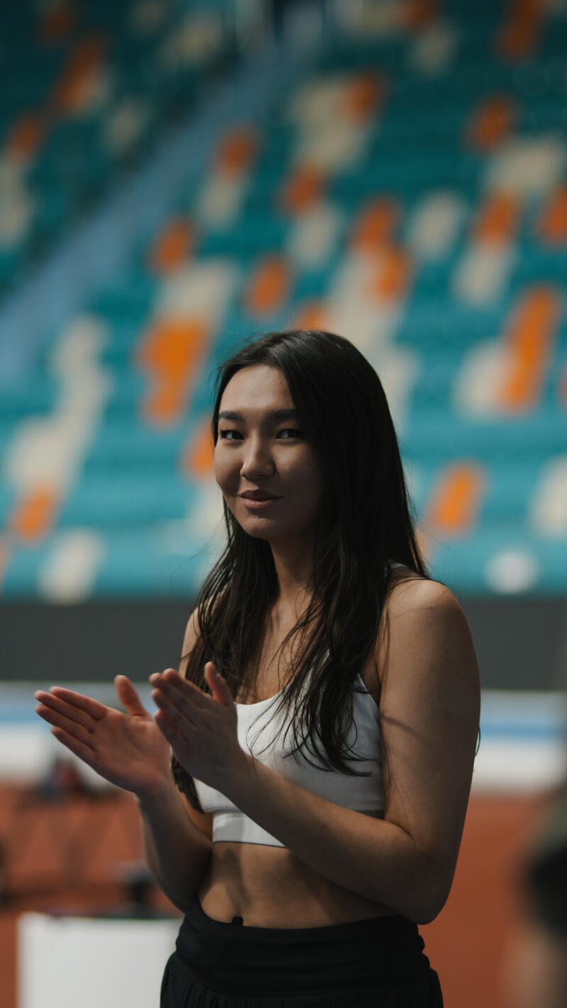 A young woman with long, dark hair, wearing a white sports bra and black pants, is clapping her hands in front of a background of teal and orange seats.