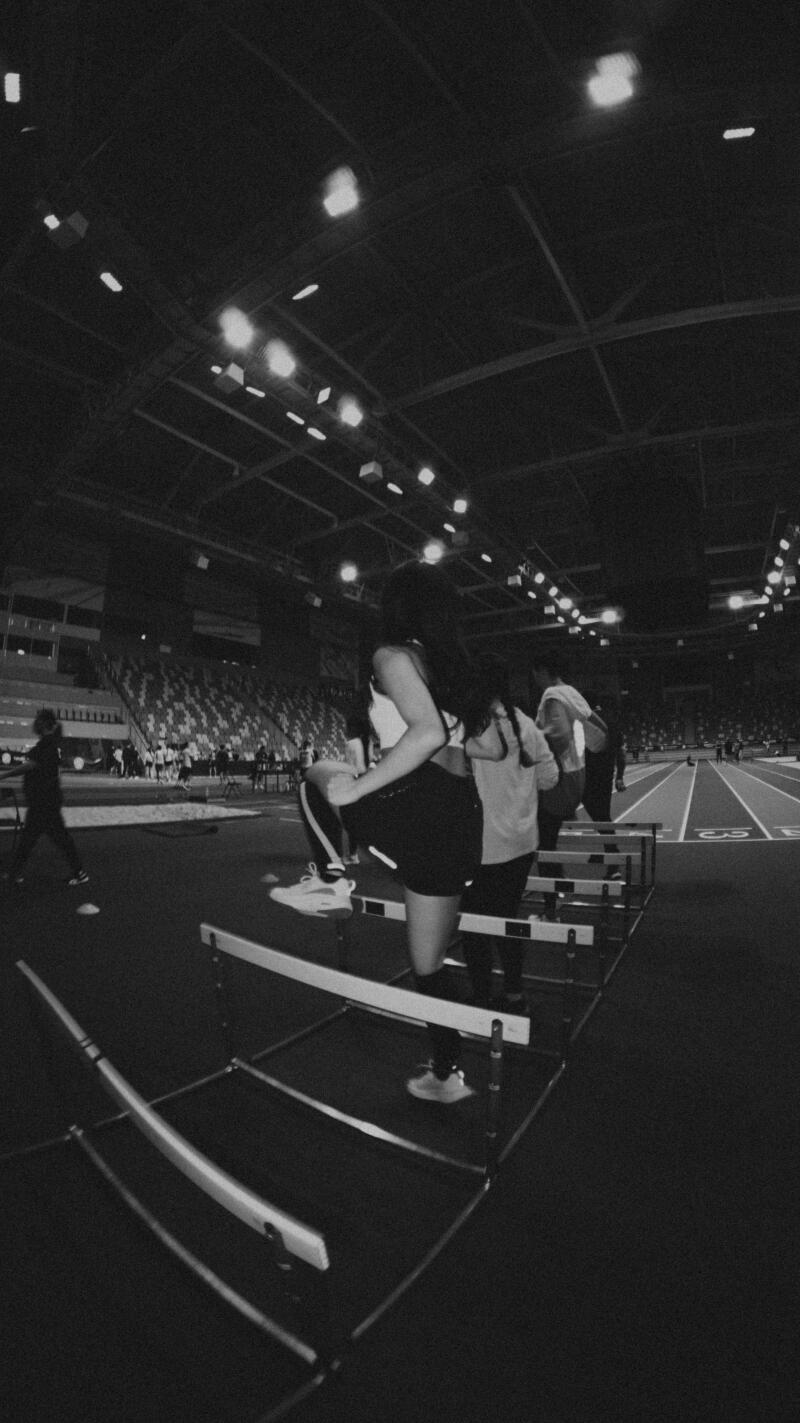 In a black-and-white shot, a woman is stepping over hurdles on an indoor track while others stand behind her.