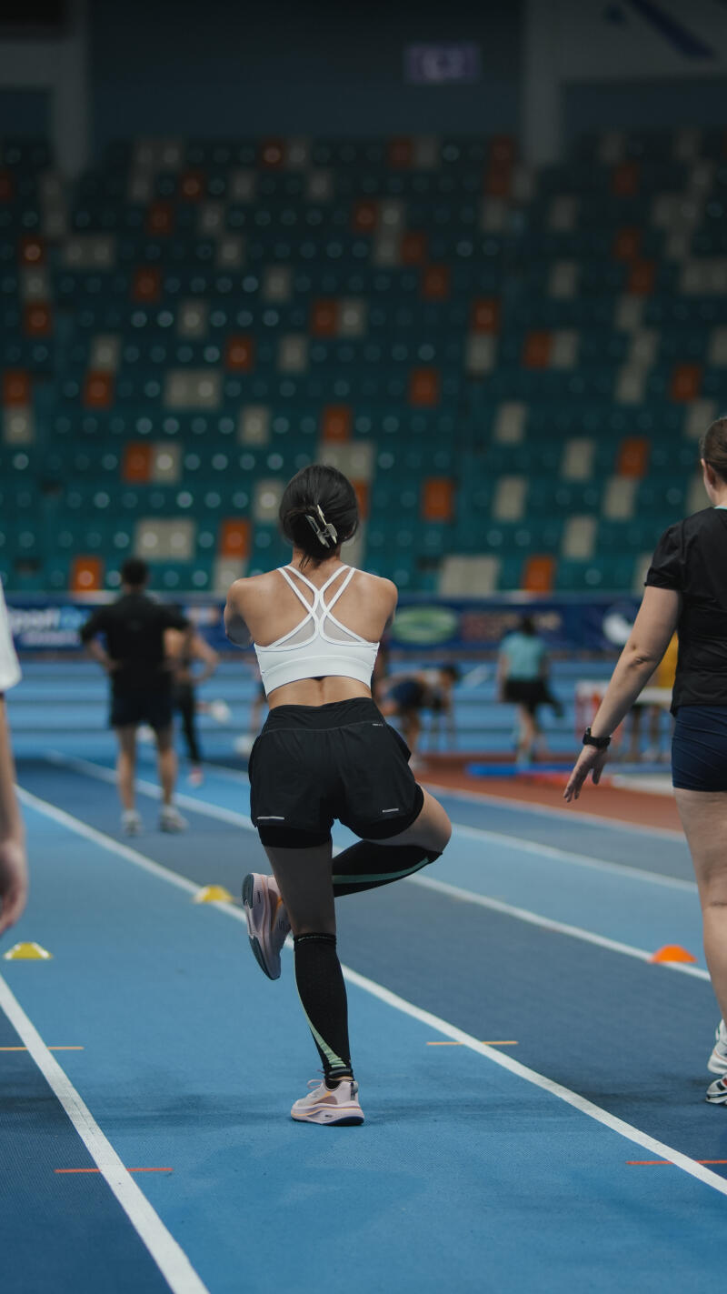 The image shows a woman in athletic wear doing a balancing exercise on a blue track, with rows of stadium seating in the background.