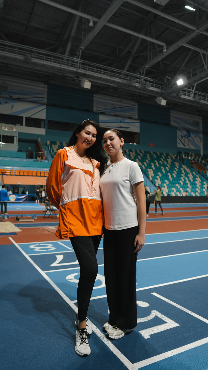 Two women stand on a blue track marked with white lines and numbers inside a stadium; one is wearing an orange and cream jacket with black leggings, while the other is in a white t-shirt and black pants.
