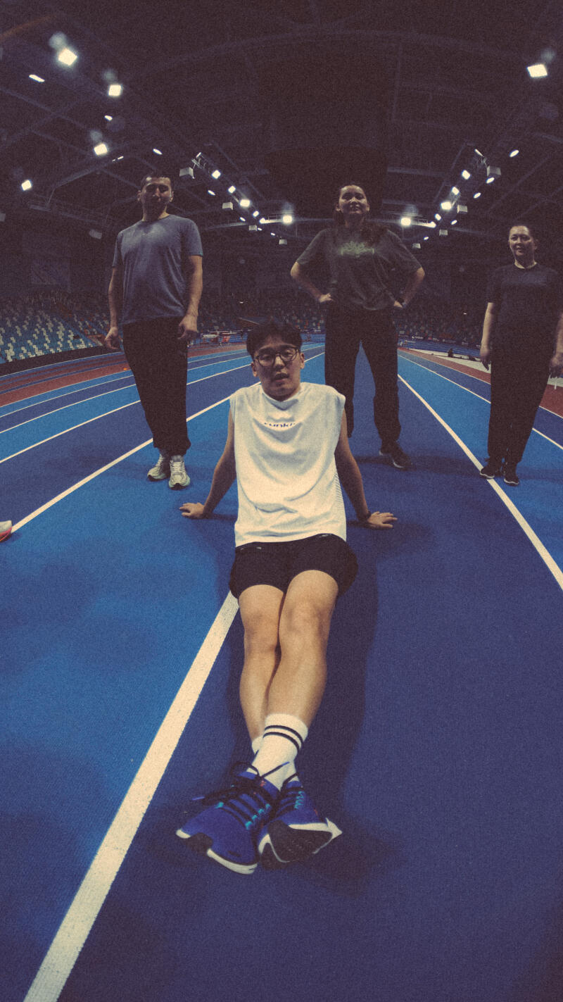 A fisheye lens captures a man sitting on a blue track with lanes while three people stand behind him in the background.