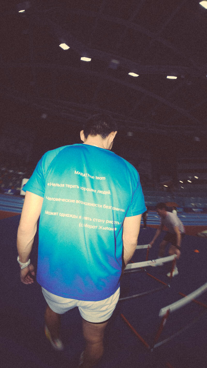 A man in a turquoise t-shirt with the text "MARAThon team", "Нельзя терять хороших людей.", "Человеческие возможности безграничны", "Может однажды я пять стону рисять (с) Марат Жылан в" is standing near hurdles.