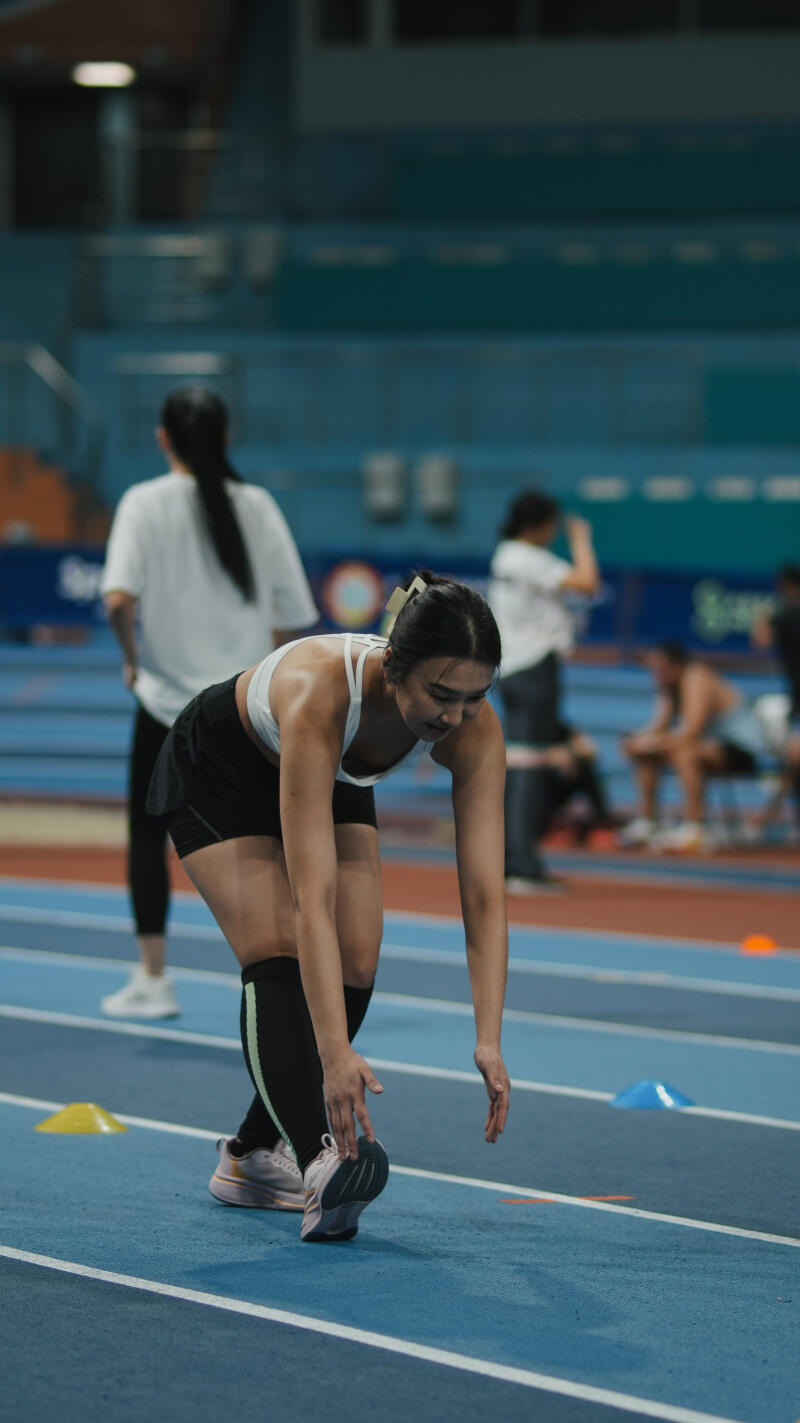 On a blue running track, a woman in sportswear bends forward to stretch, with other people visible in the background.
