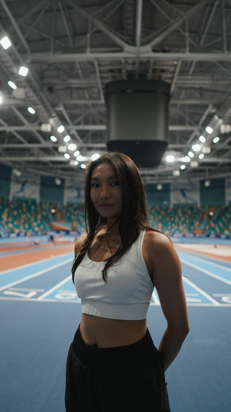 A woman in a white top and black pants stands on an indoor running track, with stadium seating and overhead lights in the background.