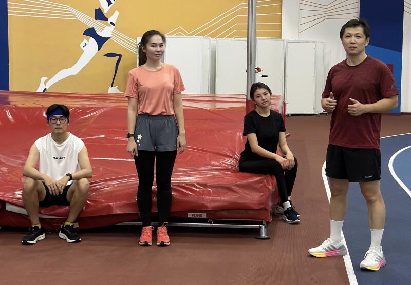 Four people are posing in a gymnasium with a red mat, including a man giving a thumbs-up while wearing a maroon shirt and black shorts.