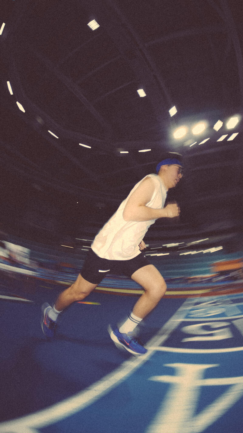 A man in a white tank top, black shorts, and blue running shoes is running on a blue track in a stadium with lights above.